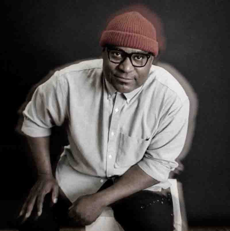 Ein Porträt von André Mack vor einem dunklen Hintergrund. Er trägt eine auffällige rote Strickmütze, eine schwarze Brille mit dickem Rahmen und ein hellgraues Hemd.