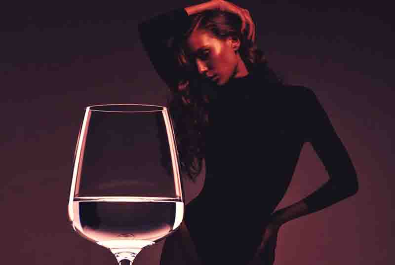 Eine kunstvolle Studioaufnahme in warmen Rosa- und Lilatönen. Im Vordergrund steht ein großes, bauchiges Weinglas, das teilweise mit einer klaren Flüssigkeit gefüllt ist; die Lichtbrechung erzeugt ein leuchtendes horizontales Band im Wasser.