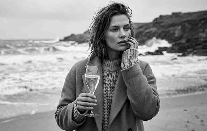 Eine Schwarz-Weiß-Fotografie einer Frau am Strand bei bewölktem Wetter. Sie trägt einen dicken, grob gestrickten Pullover und einen schweren Mantel. In der einen Hand hält sie ein Sensorik Champagner-Verkostungsglas.