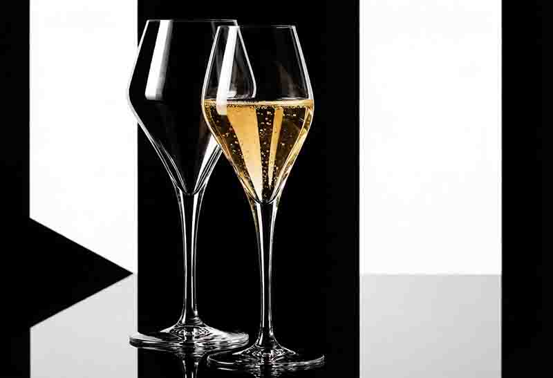 Zwei Champagner Sensorik Tastinggläser vor einem kontrastreichen schwarz-weißen Hintergrund zeigt. Eines der Gläser ist mit Champagner gefüllt, der feine Blasen aufweist.