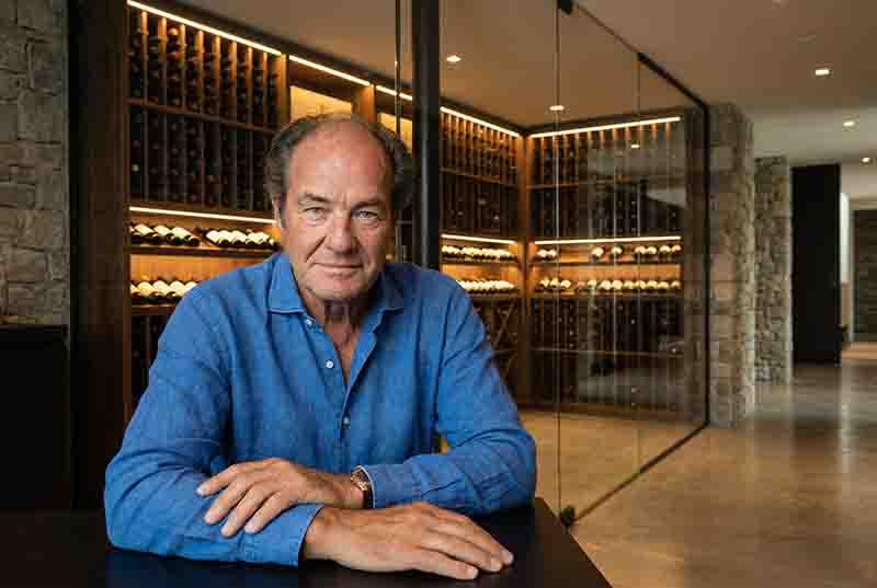 Porträt Georg Josef Riedel,, der an einem dunklen Tisch sitzt. Im Hintergrund ein moderner, Weinkeller mit Glaswänden.