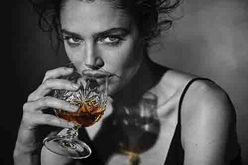Schwarz-Weiß-Fotografie einer Frau, die ein Kristallglas mit einem bernsteinfarbenen Getränk hält.
