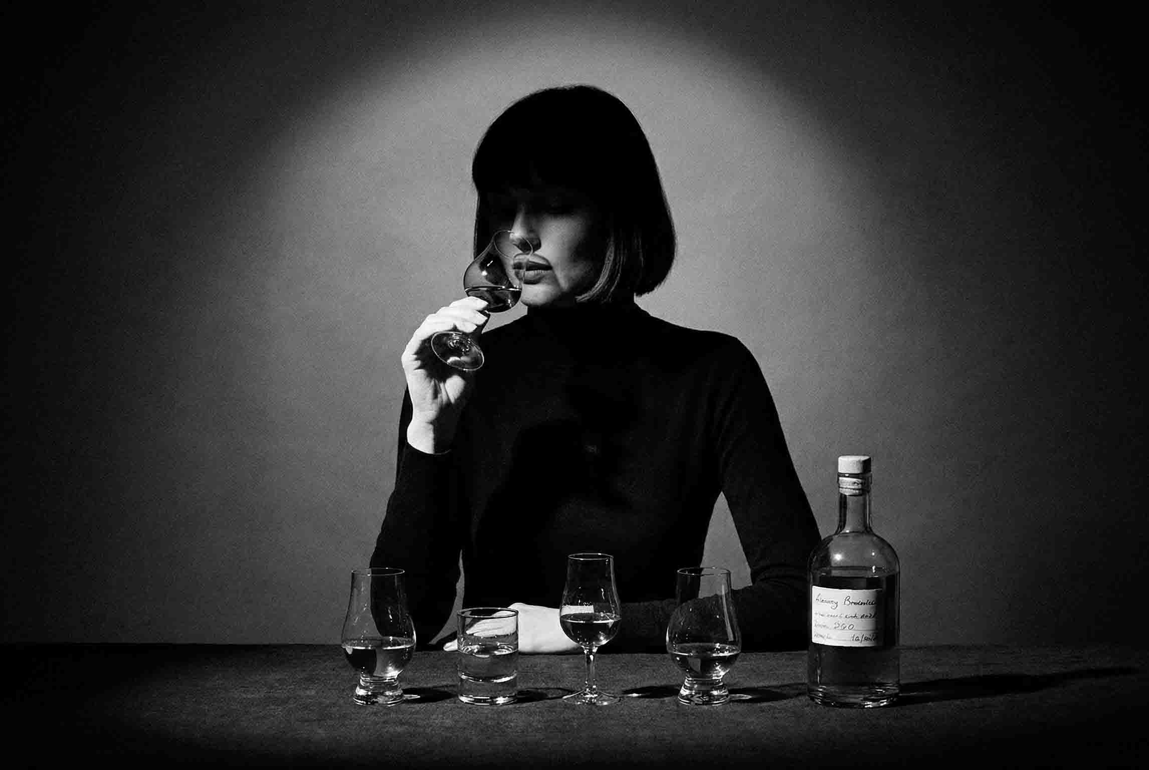 Schwarz-Weiß-Fotografie einer Frau mit dunklem Pagenkopf und schwarzem Rollkragenpullover. Sie riecht konzentriert an einem Degustationsglas (Glencairn-Stil). Vor ihr auf einem dunklen Tisch stehen vier verschiedene Probiergläser sowie eine klare Glasflasche mit einem handbeschriebenen Etikett. Ein starkes Spotlight von oben erzeugt eine stimmungsvolle Noir-Atmosphäre mit tiefen Schatten.