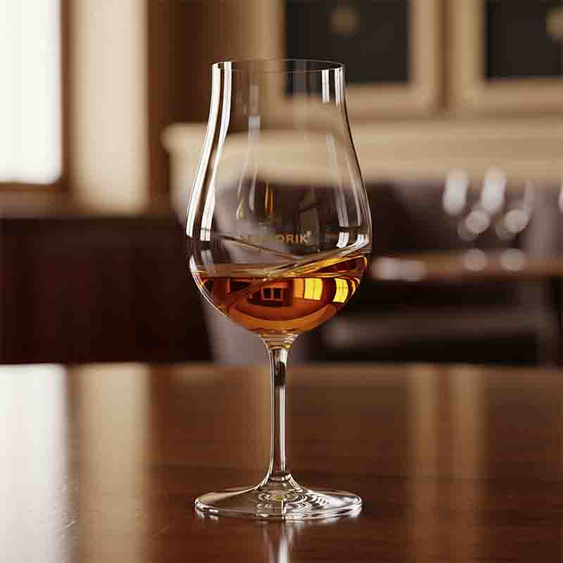 Ein Nahaufnahme-Foto eines eleganten, tulpenförmigen Sensorikglas/Degustationsglas, das teilweise mit einer bernsteinfarbenen Flüssigkeit (vermutlich Whisky oder Cognac) gefüllt ist.