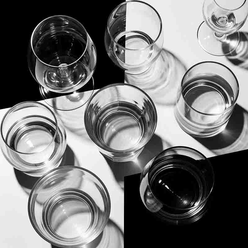 Schwarz-Weiß-Foto in Aufsicht und hohem Kontrast, das sieben klare Gläser und Kelche zeigt, von denen einige mit Wasser gefüllt sind. Sie stehen auf einer Oberfläche, die in schwarze und weiße Quadrate unterteilt ist. Starke Schatten werden von den Glaswaren geworfen und erzeugen abstrakte Muster und Reflexionen.