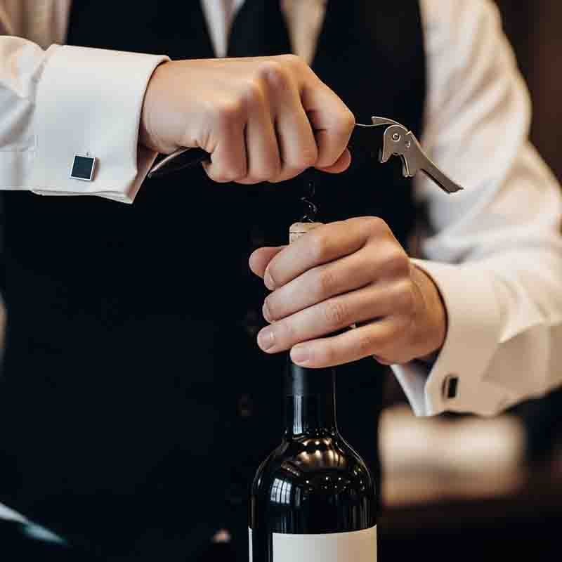 Eine Nahaufnahme der Hände eines Sommeliers, die ein weißes Hemd mit Manschettenknöpfen und eine dunkle Weste trägt und einen Korkenzieher benutzt, um eine Flasche Wein zu öffnen.